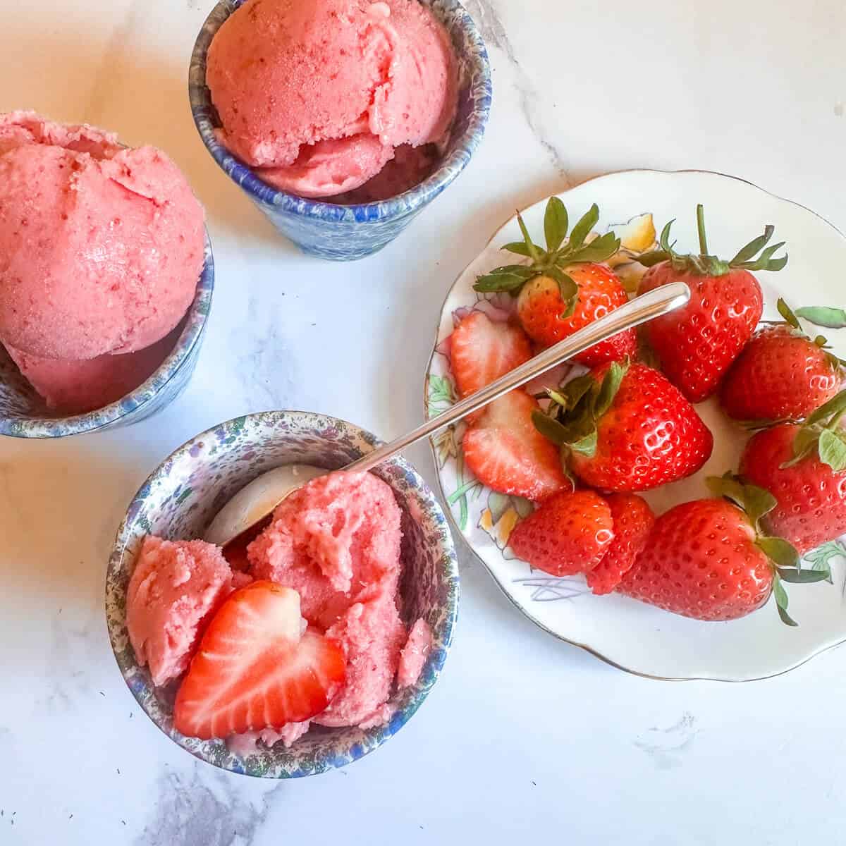 A plate of strawberries next to 3 bowls of strawberry frozen yogurt.