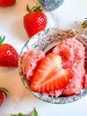 A small blue bowl with a scoop of strawberry frozen yogurt in it, topped with a slice of strawberry and surrounded by fresh strawberries.
