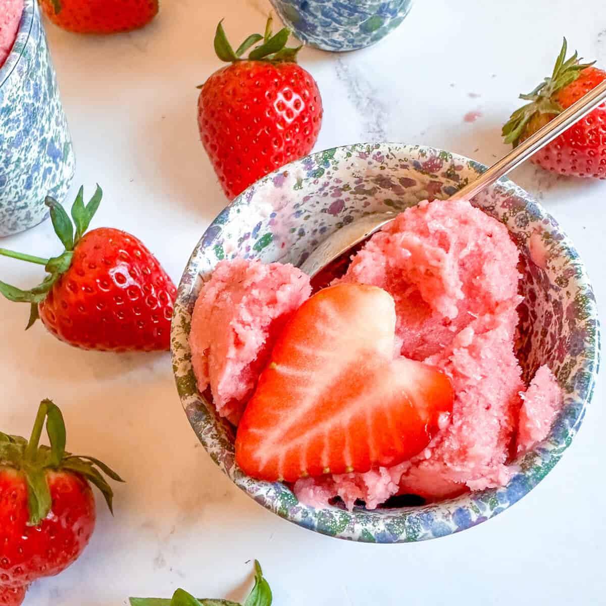 A small blue bowl with a scoop of strawberry frozen yogurt in it, topped with a slice of strawberry and surrounded by fresh strawberries.