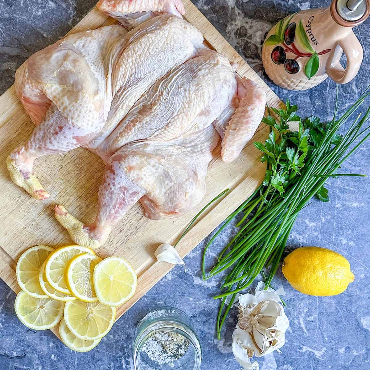 A spatchcock chicken on a chopping board next to some lemons, olive oil, garlic cloves and fresh herbs. 