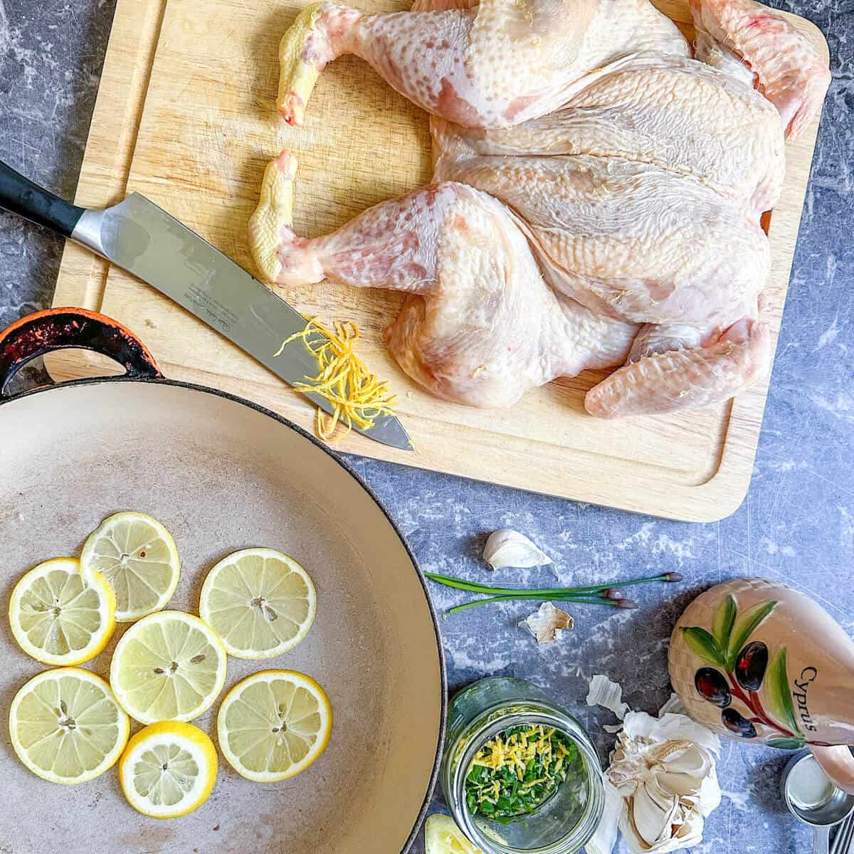 A shallow cast iron pan with slices of lemon in the base. Next to it, a raw spatchcock chicken is on a chopping board next to the other ingredients for the marinade. 