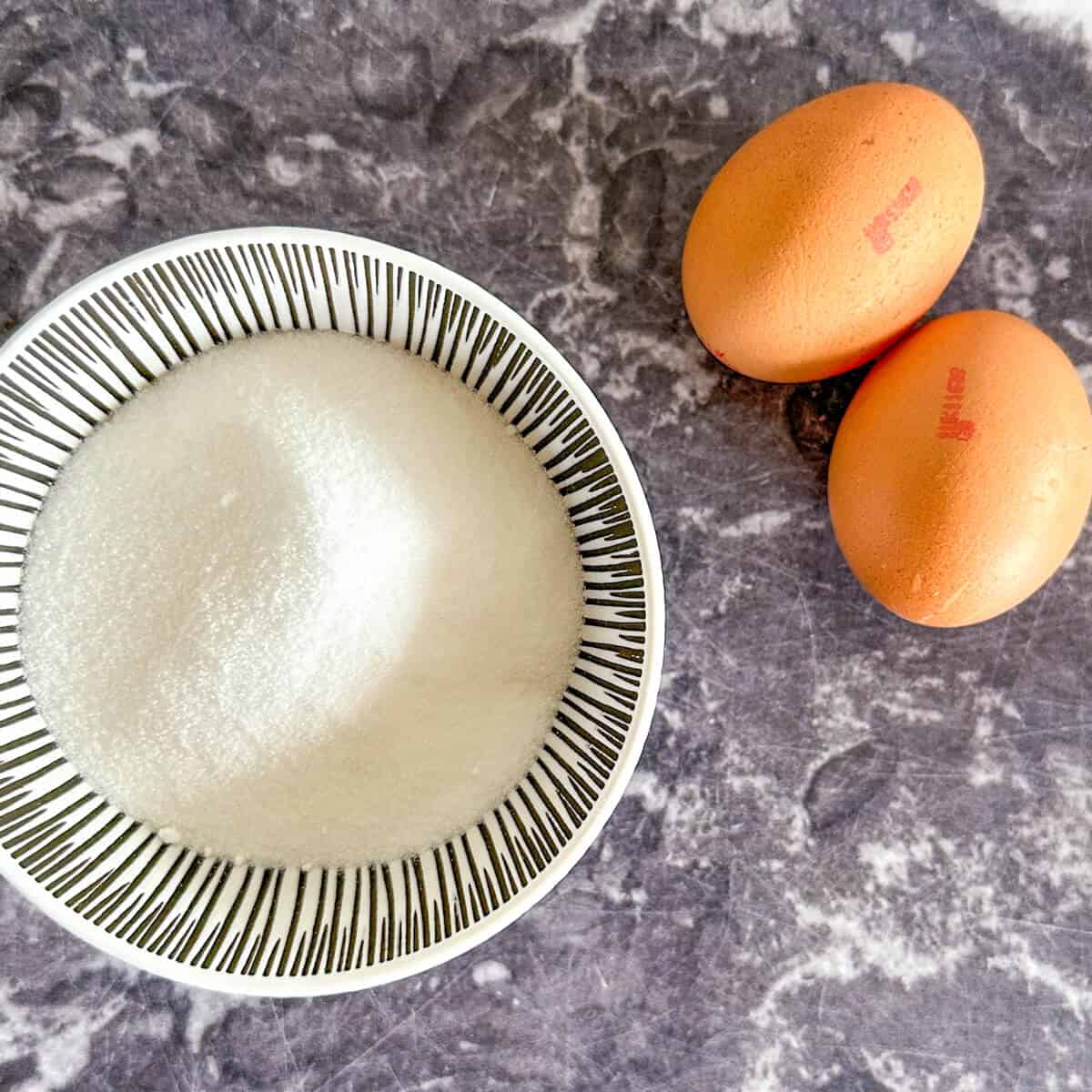 A bowl of sugar next to two eggs. 