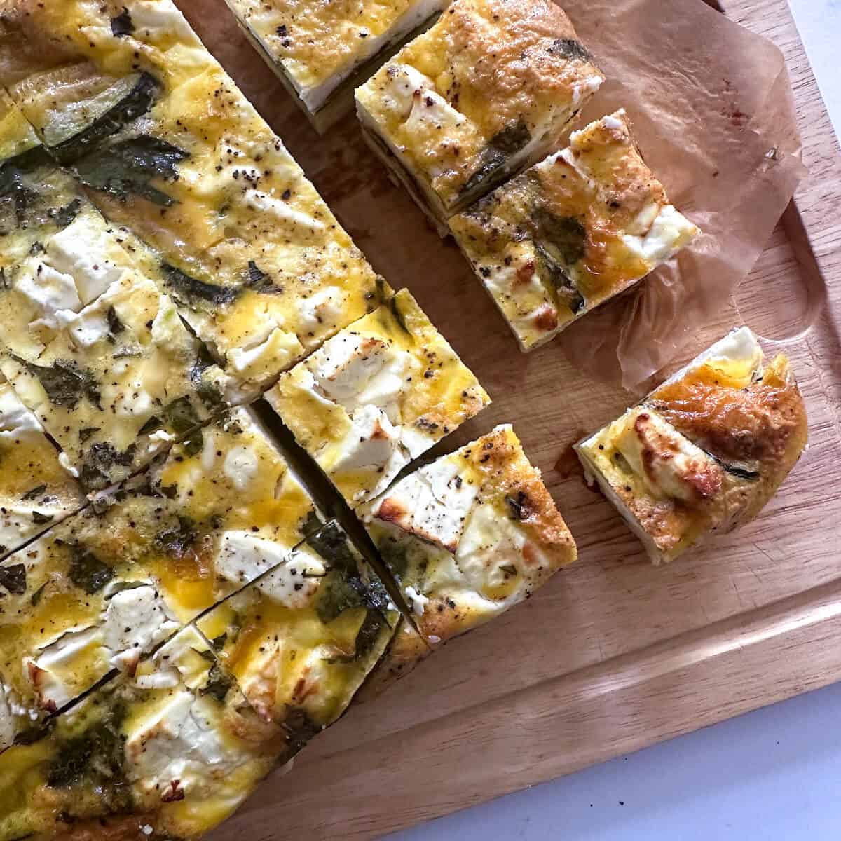 A wooden serving board with a baked frittata on it cut into small squares.