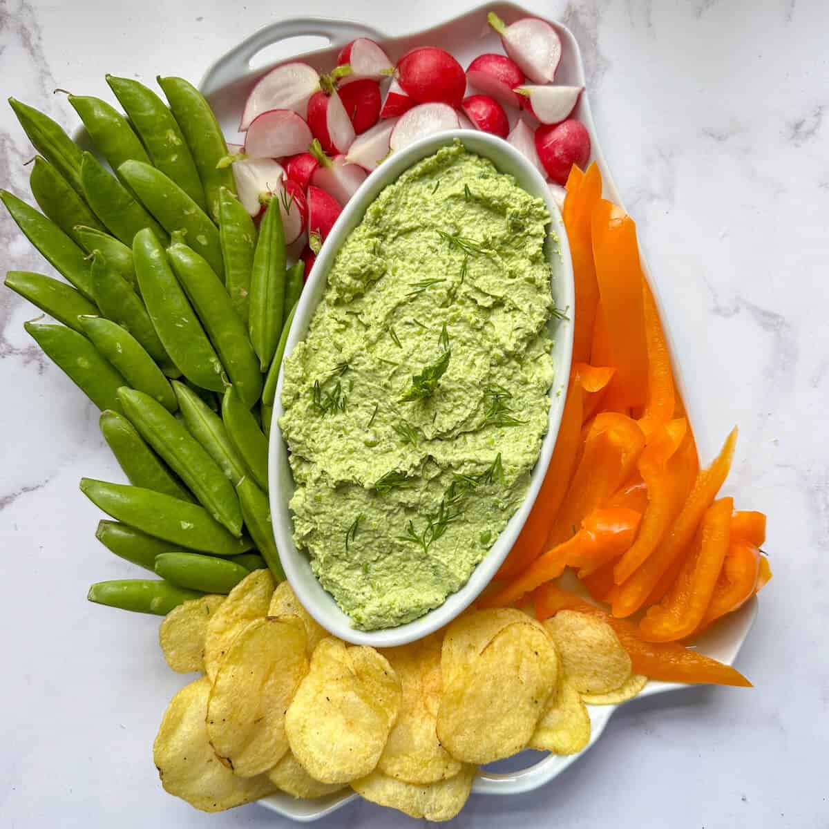 A bowl of green pea dip surrounded by colorful vegetable crudites and crisps.