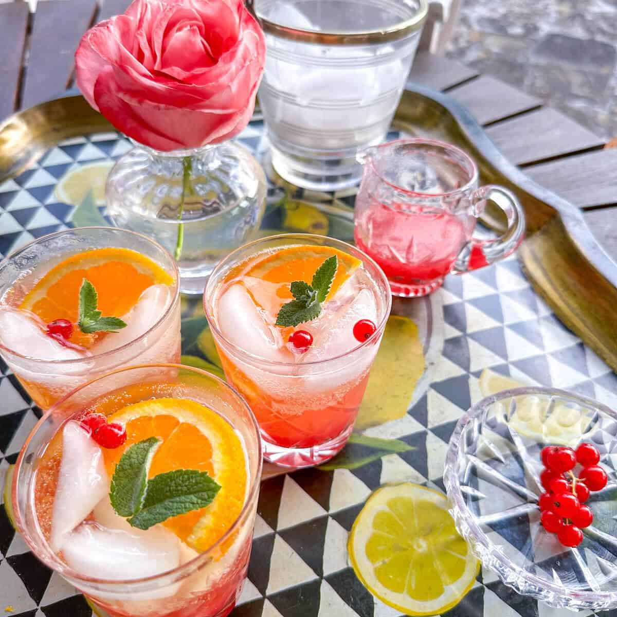 A serving tray with 3 glasses of red currant cordial drinks next to a small jug of cordial, an ice bucket and a vase with a rose in it. 