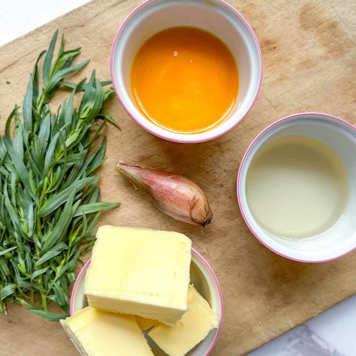 The ingredients for bearnaise sauce: Fresh tarragon next to pots of egg yolks, white wine vinegar, butter and a shallot on a wooden chopping board. 