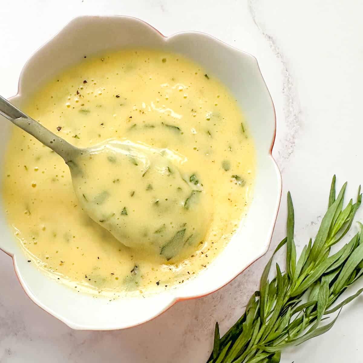 A dish of bearnaise sauce with a spoon in it. Next to a small bunch of fresh tarragon. 