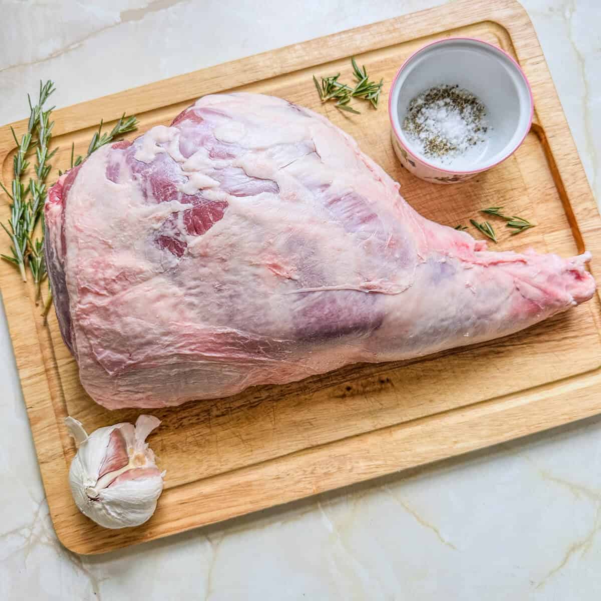 A raw leg of lamb on a wooden chopping board next to some sprigs of rosemary, a pot of salt and pepper, and garlic cloves. 