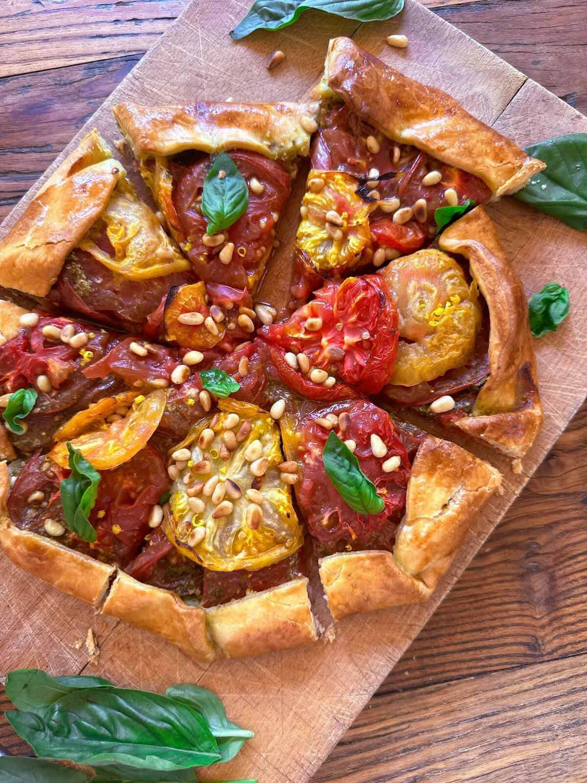 A fresh tomato galette on a wooden chopping board with pine nuts and basil leaves scattered on top. 