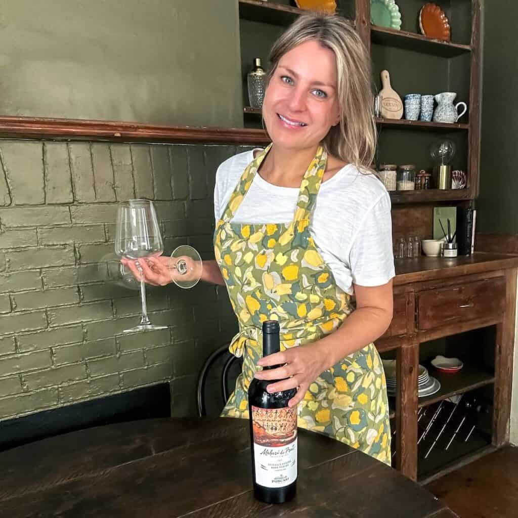 Rosanna Stevens, founder of Rosanna ETC standing in a kitchen holding a bottle of wine and two wine glasses.