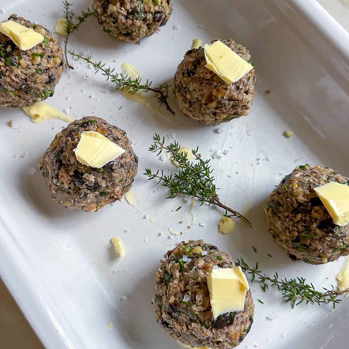 An oven dish with six mushroom stuffing balls ready to be baked, each topped with a slice of butter and some fresh thyme. 