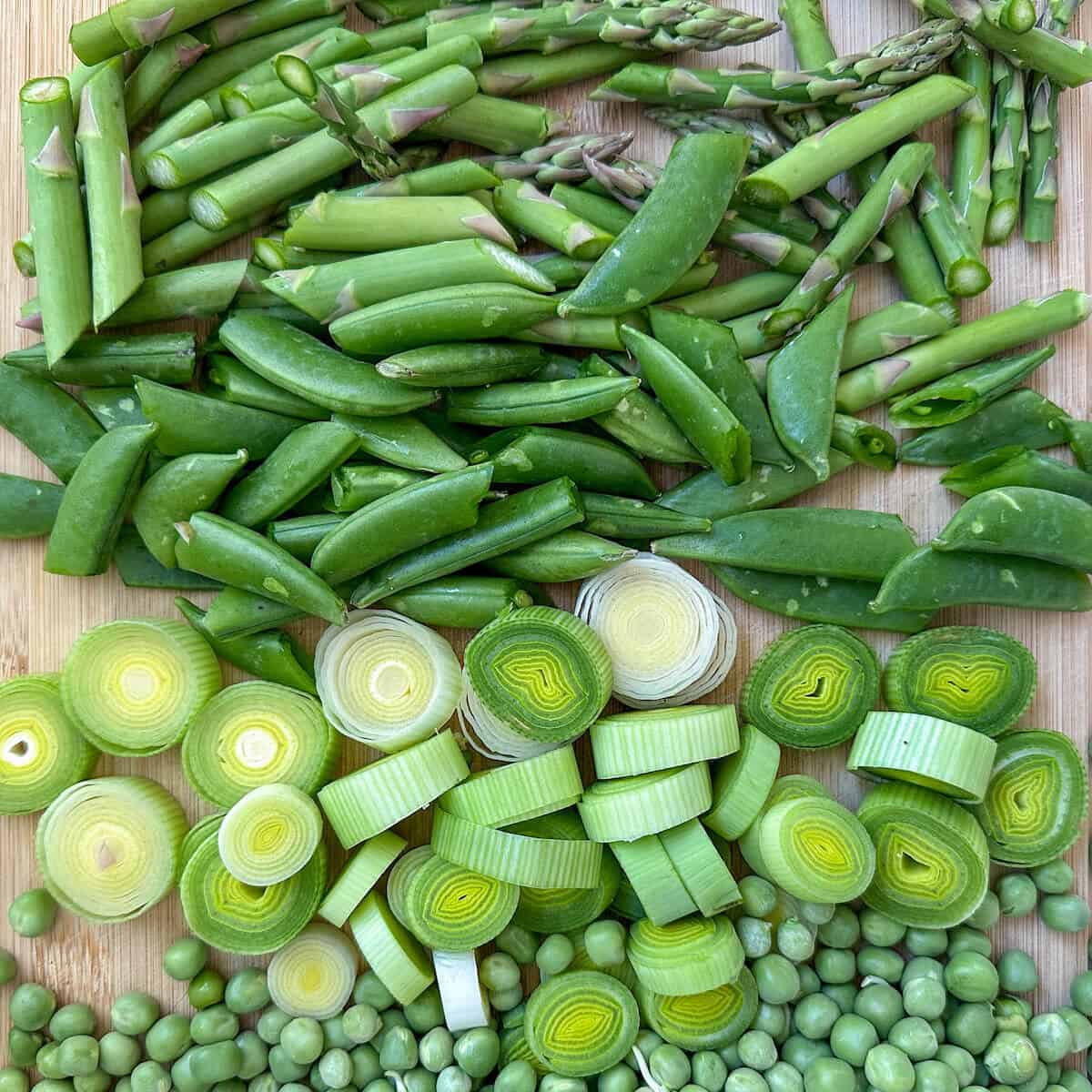 A chopping board with asparagus, peas, sugar snap peas and leeks on it.