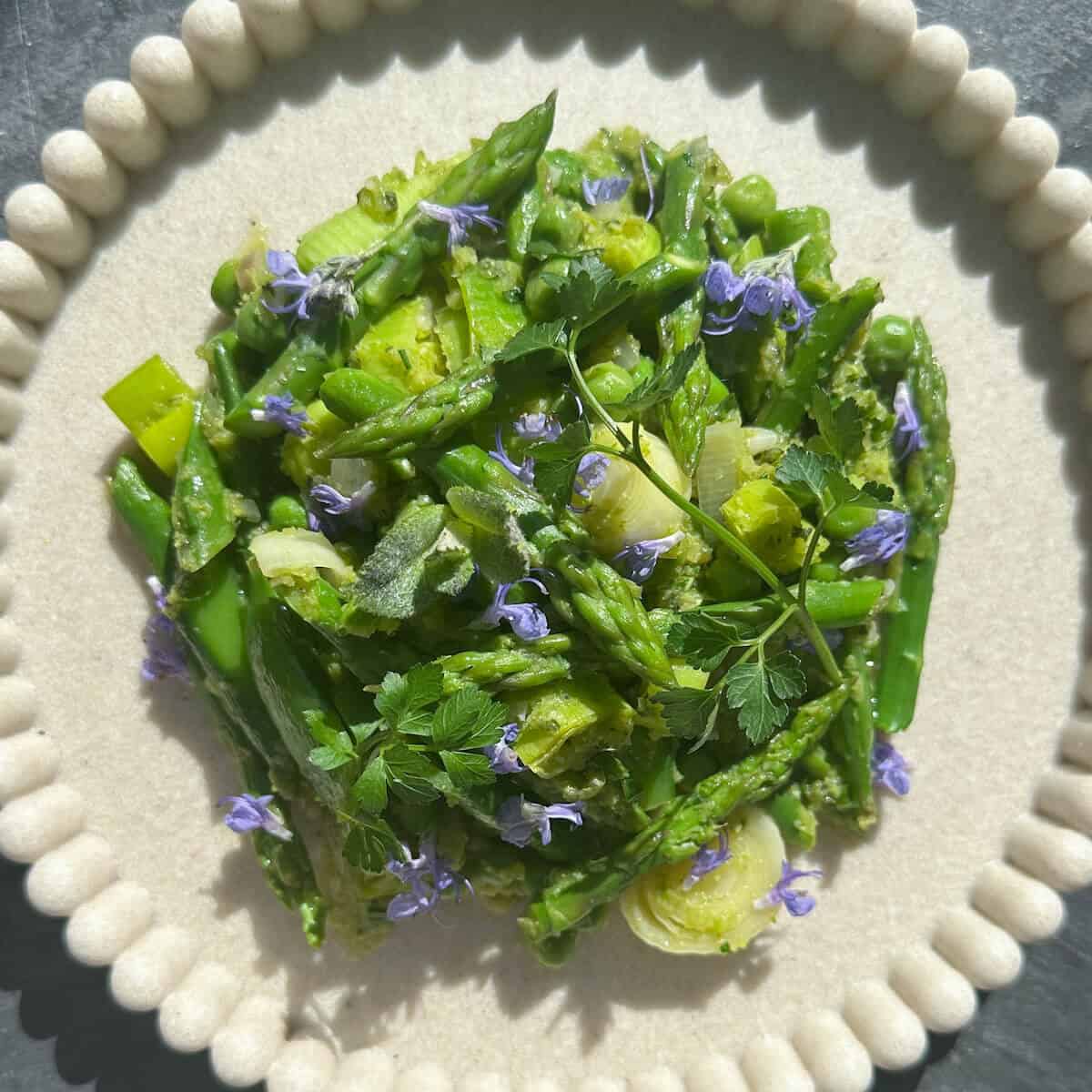 A plate in sunny sunshine with a vibrant green spring vegetable fricassee on it and purple edible flowers.