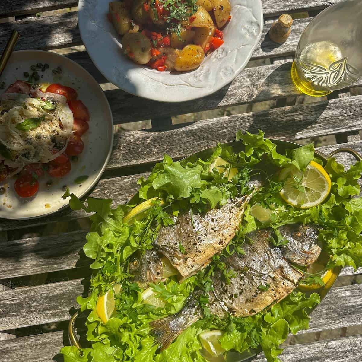 A table of dishes, including whole baked sea bream, fennel tomato salad, and potato salad. 